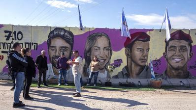 Eine Gruppe steht vor einer Betonmauer. Auf der Mauer sind farbige Portraits von israelischen Geiseln zu sehen.