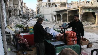A vegetable stall in the middle of ruins, a elderly woman sits on a chair, two men are selling/buying.