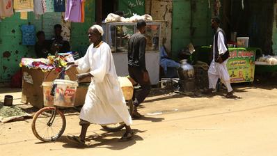 A man pushes his bicycle along a sandy path. In the background a green wall, several people walking and clothes being hung up to dry.
