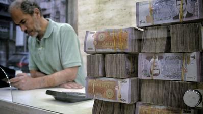 A man stands at a desk looking at his phone. In the foreground, a stack of 2000 SYP notes bearing the face of Bashar al-Assad.