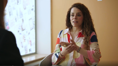 Eine Frau in bunter Bluse sitzt vor einem Fenster und spricht mit ihrem Gegenüber.