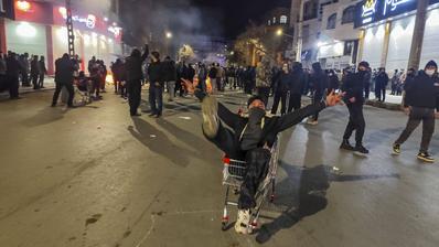 A man wearing a lower face covering sitting in a shopping trolley. Around him are other people protesting.