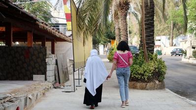 Zwei Frauen laufen in einer Straße und halten Hände. Eine trägt ein Kopftuch, die andere nicht.  