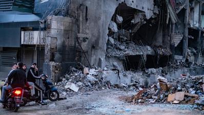 A group on motorbikes in the foreground on the left. In the background a bombed building and rubble.