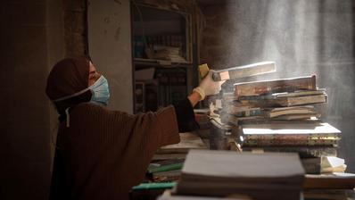 Eine Frau mit Hijab und Atemmaske entstaubt Bücher in einer zerstörten Bibliothek.