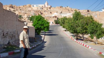A man stands beside a road with a hilltop village in the background.