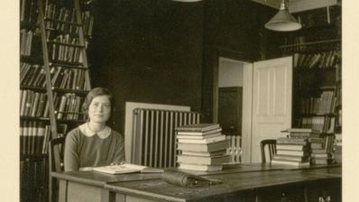 A black and white image of a young woman sitting in a room full of books.