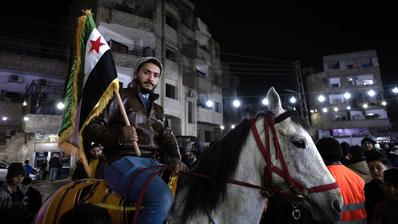 Ein Mann sitzt auf einem weißen Pferd und hält die neue syrische Flagge. Hinter ihm Gebäude in der Nacht.