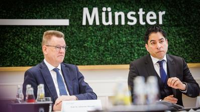 Zwei Männer in Anzug sitzen an einem Konferenztisch, im Hintergrund an der Wand der Schriftzug "Münster"