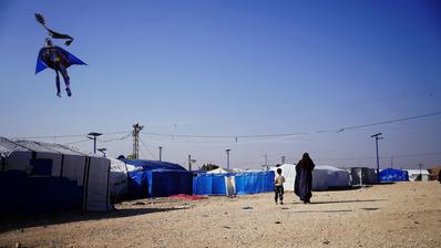 A woman and a child walk towards a cluster of tents. A Batman shaped kite is visible in the sky.