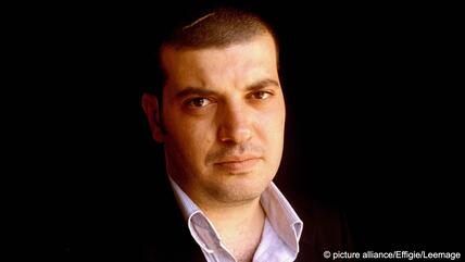 Headshot of Palestinian writer Sayed Kashua wearing an open-necked shirt and jacket against a dark background