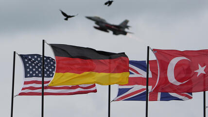 American, German, English and Turkish flags are waving in the wind. A NATO fighter jet flies above them.