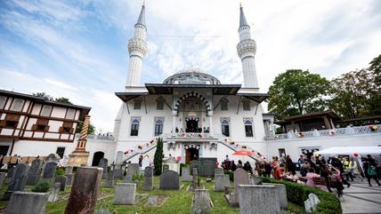 People gather outside a white mosque with colourful balloons attached to the fence. In the foreground is a graveyard.