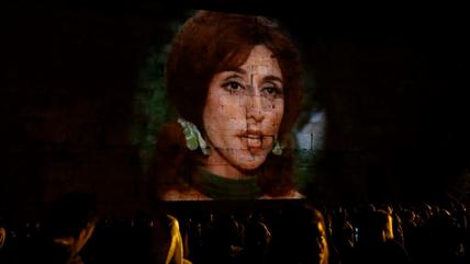 The face of a woman is projected onto a wall made of large stone blocks.