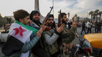 A group of young men carrying guns, one has a Syrian revolutionary flag wrapped around his shoulders.