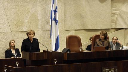Angela merkel stands at a podium speaking into a microphone. An Israeli flag hangs in the background.