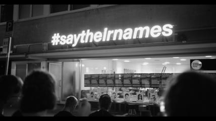 B/W photo of a group of people standing in front of a lit-up sign that says "Saytheirnames"