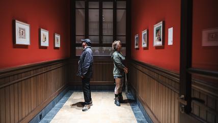 18th Istanbul Biennial (Photo: IKSV | Mete Kaan Özdilek) Two people in a corridor, facing away from eachother and looking at paintings hanging on the walls.