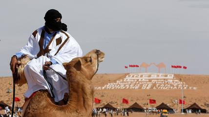 A man in Sahrawi clothing with a headscarf rides a camel. In the background, a UNESCO advertisement on a dune.