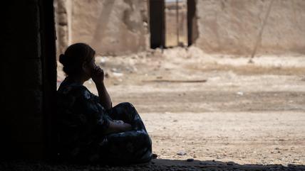 A silhouetted woman sits in the doorway of a ruined building looking out onto the street.