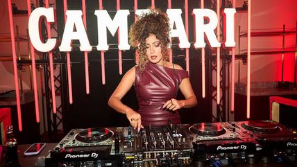 Eine Frau in dunkelrotem Kleid und hochgesteckten Haaren steht am DJ Pult. Hinter ihr Leuchtreklame.