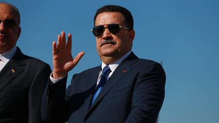 A man in a suit and sunglasses raises his hand to wave at people.