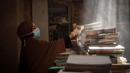 Eine Frau mit Hijab und Atemmaske entstaubt Bücher in einer zerstörten Bibliothek.