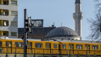 Stadtbild, im Vordergrund fährt eine gelbe Bahn, im Hintergrund ein Wohngebäude und eine Moschee.