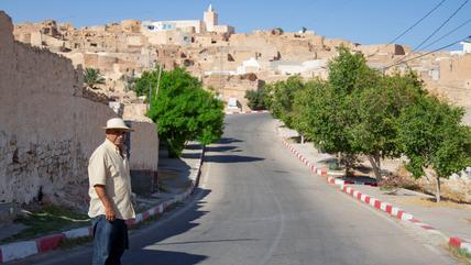A man stands beside a road with a hilltop village in the background.
