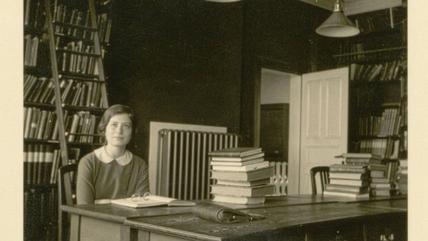 A black and white image of a young woman sitting in a room full of books.