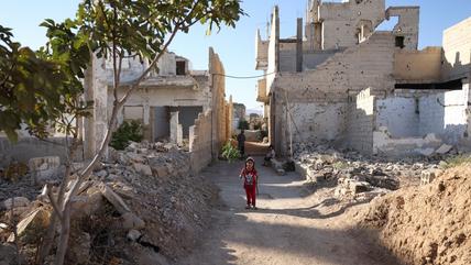 A girl is standing on a path between dilapidated buildings and rubble. In the background are another girl and an old man.