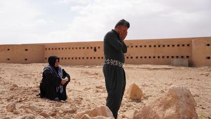 A man holds his head in his hands. Behind him a woman is sitting on the ground. In the background there is a large prison building.