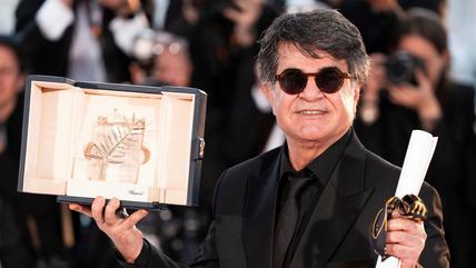 A man wearing a black suit and sunglasses holds an award and a scroll.