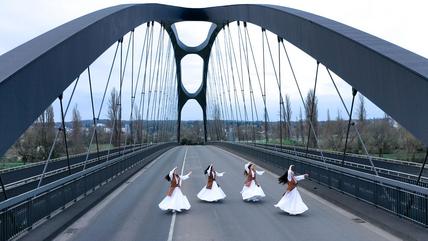 Auf einer bogenförmigen Autobahnbrücke tanzen vier Frauen in weißen Gewändern und langen dunklen Haaren.