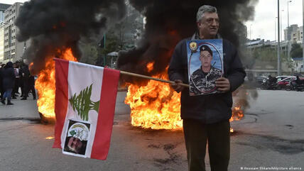 رجل واقف بجانب نار في أحد شوارع بيروت حاملاًَ العلم اللبناني وصورة جندي.