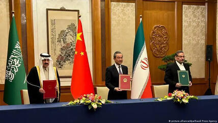 ثلاثة رجال يقفون خلف طاولة ويحملون وثائق - اتفاق إيراني سعودي برعاية صينية.
