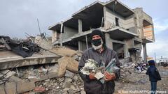 Masked man clasps tattered pile of books in front of the ruins of a building against a grey sky