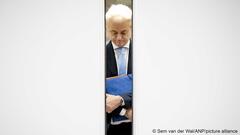Geert Wilders, holding a blue folder and looking at the ground, can be seen through a crack in an open door 