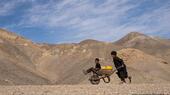 An Afghan boy pushes a wheelbarrow with canisters and his younger brother, on their way to collect water. Mountains can be seen in the background