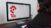 Woman sits in front of a screen displaying a cross-stitch pattern of a red flower