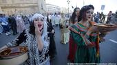 Women in traditional costumes and carrying large bowls and containers walk down the middle of a street as part of a protest on the 61st anniversary of Algerian independence while people on the footpaths and police officers look on