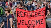 Two men carry a red banner reading "Refugees Welcome" through a crowd of people