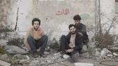 Three members of El Khat sit on the ground and on pieces of rubble in front of a white wall.