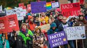 A crowd holding signs and banners demonstrate against the far right.