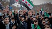 A crowd of people make peace signs to the camera. Syrian revolutionary flags wave in the background.