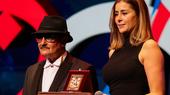 A man in a black hat and a woman stand behind a podium. She is holding a microphone.