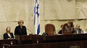 Angela merkel stands at a podium speaking into a microphone. An Israeli flag hangs in the background.