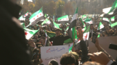 A crowd of people celebrating, many Syrian revolutionary flags are being waved.