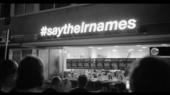 B/W photo of a group of people standing in front of a lit-up sign that says "Saytheirnames"