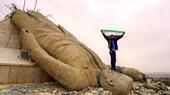 A man with the flag of free Syria stands on the head of a toppled Assad statue.